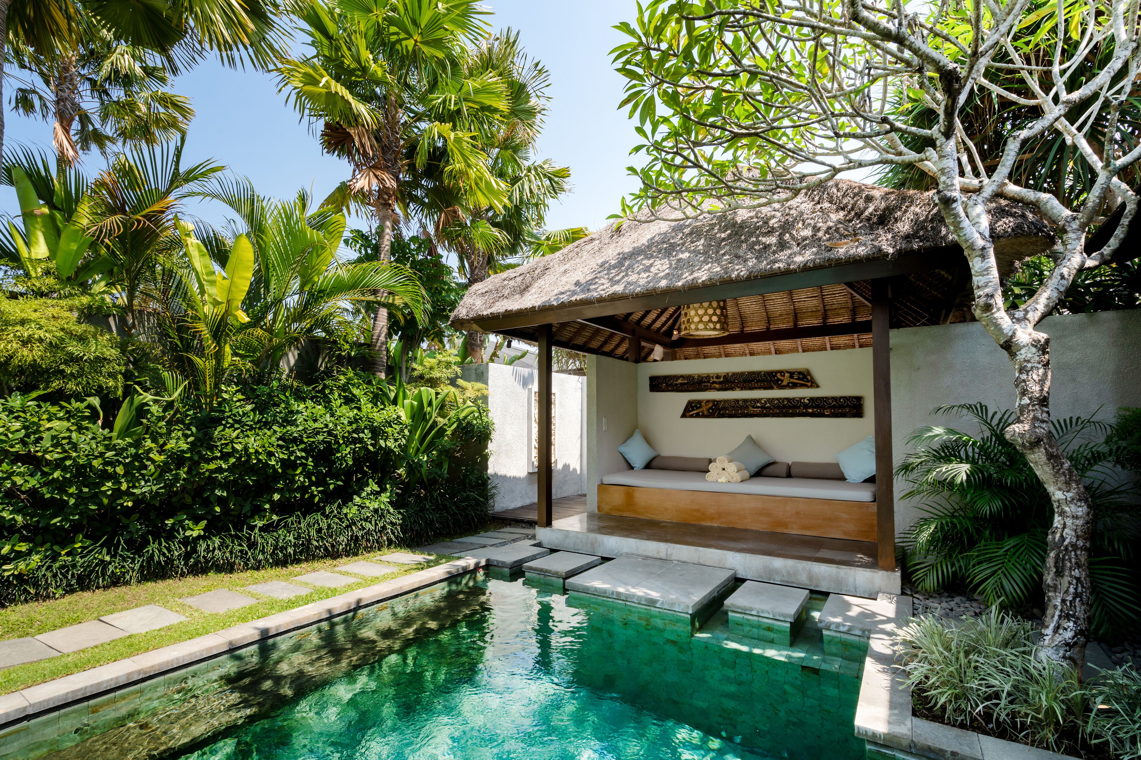 A pool area with a thatched-roof structure, a seating area with cushions, and lush greenery surrounding the space.