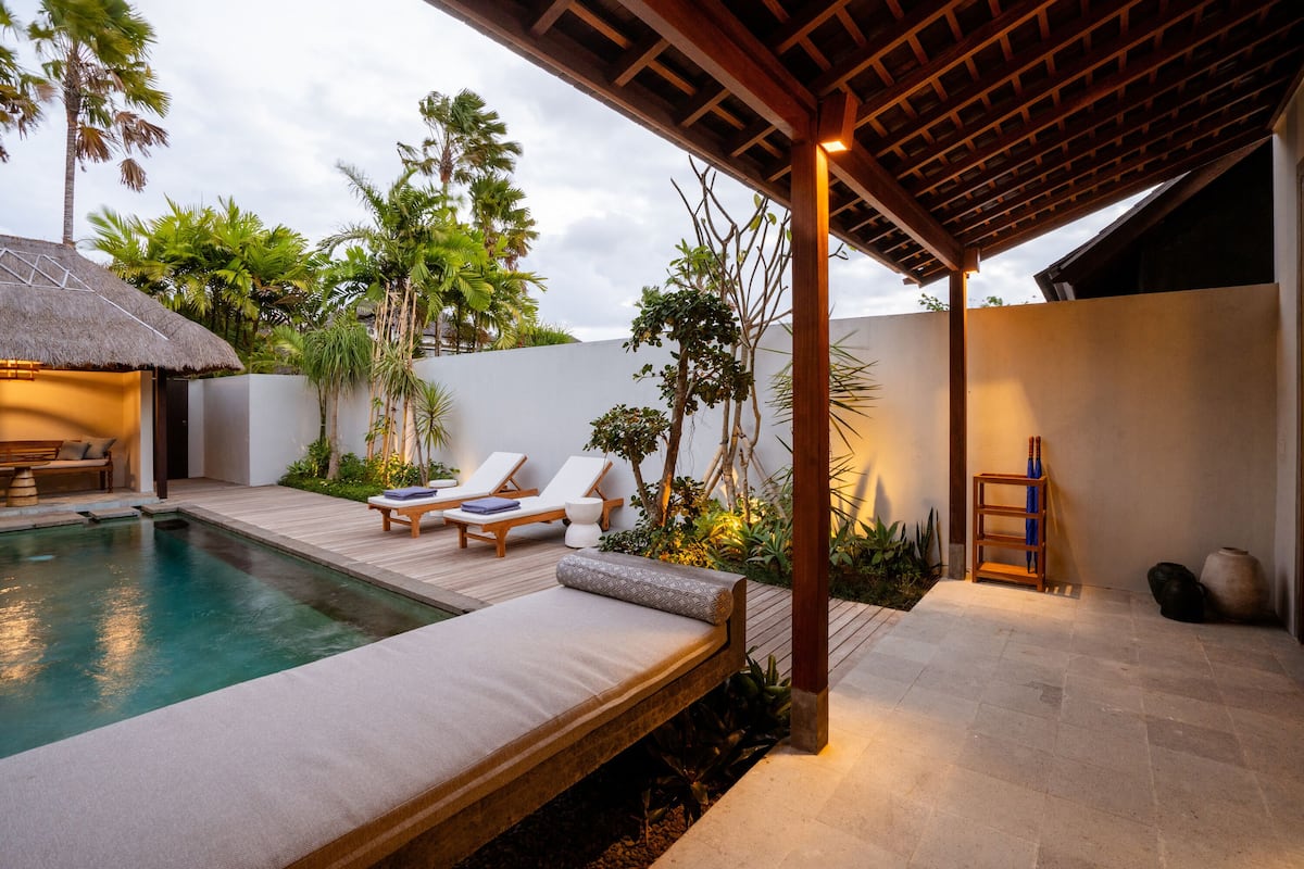 Un área junto a la piscina con una silla de descanso, una mesita y una estructura con techo de paja.