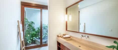 Un baño moderno con un espejo grande, armarios de madera y vistas a un jardín a través de una puerta de cristal.