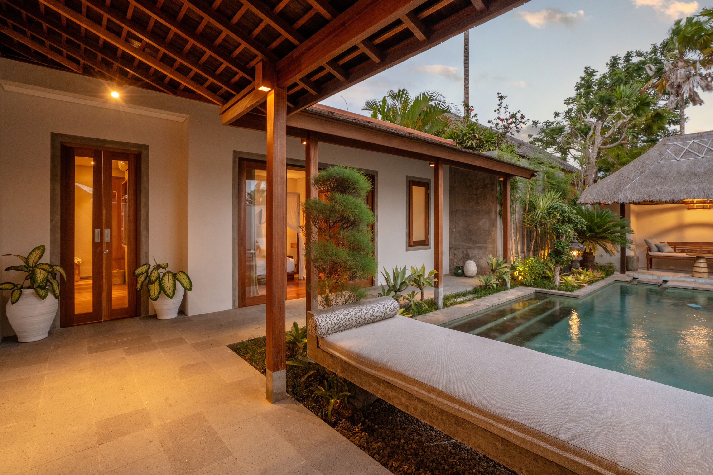 A poolside area with a lounge chair, a thatched-roof structure, and potted plants.