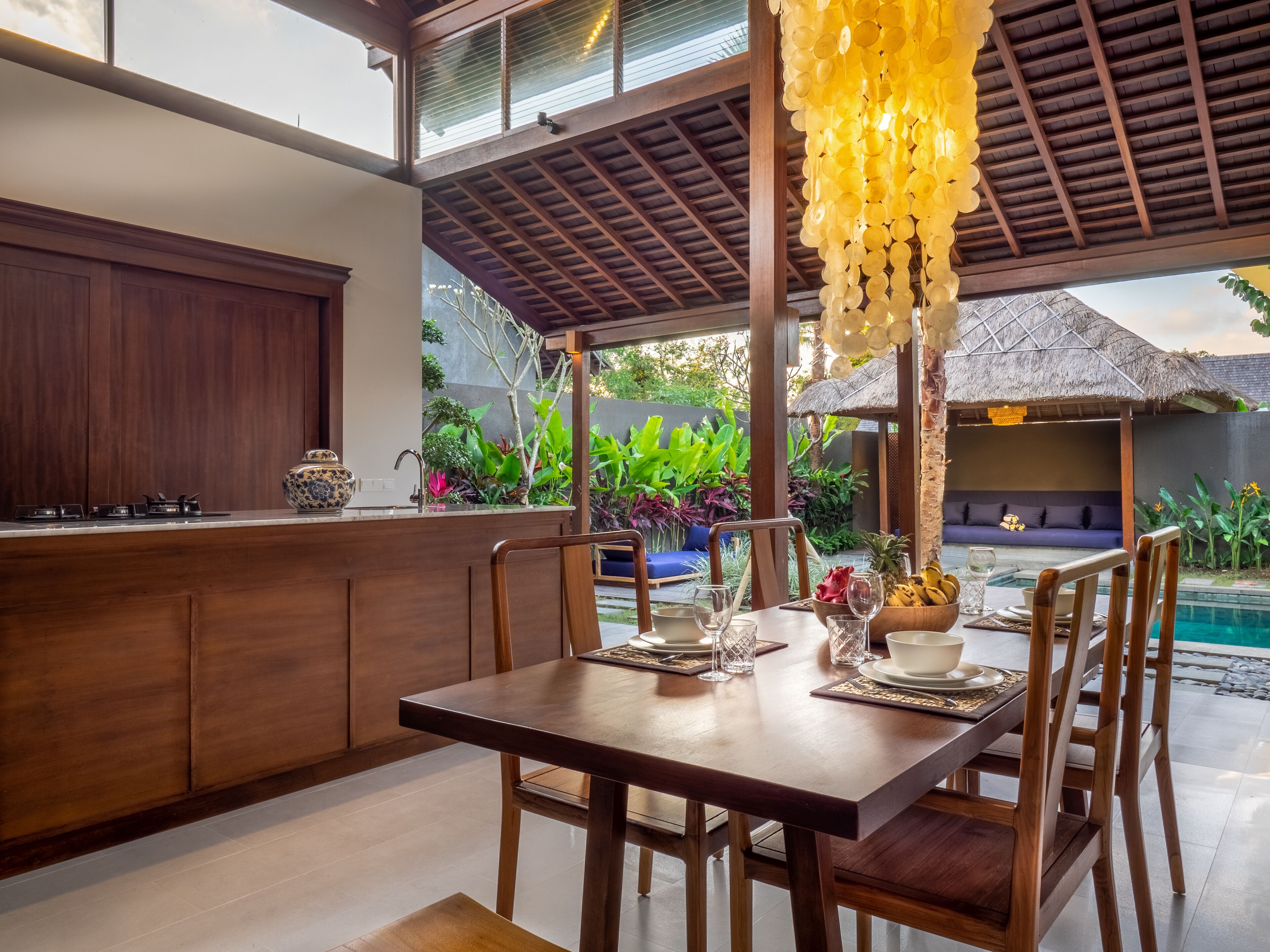 Un coin repas avec une table et des chaises en bois, une cuisine dotée d’une cuisinière intégrée et, au fond, une construction au toit de chaume.