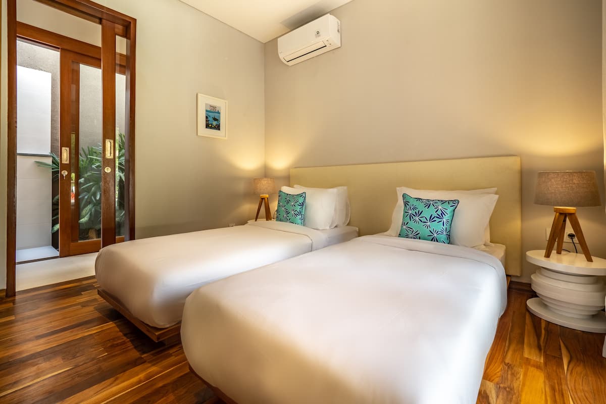 Habitación de hotel con dos camas, suelo de madera, unidad de aire acondicionado, una mesita de noche con lámpara y puerta con panel de vidrio.