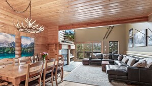 A wooden interior with a dining table, chairs, a sofa, and a large window overlooking a mountainous landscape.