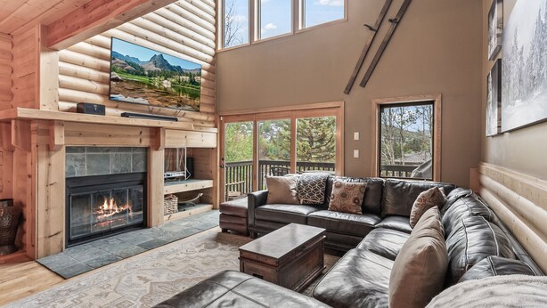 A log cabin interior with a fireplace, leather sofa, and large windows offering views of the outdoors.