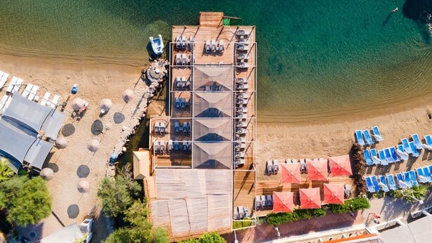 Vista aérea de un hotel frente al mar con una terraza de madera, sillas de descanso y sombrillas.