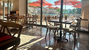 Un área de comedor interior con mesas y sillas, ventanas grandes y sombrillas rojas en el exterior.