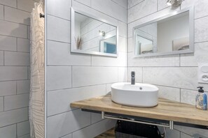 Une salle de bain moderne avec un meuble-lavabo en bois, un lavabo blanc, deux miroirs fixés au mur et un rideau de douche orné d’un motif de feuilles.