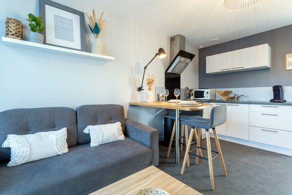 Un salon moderne avec un canapé gris, une table basse en bois et un coin cuisine doté de meubles blancs et d’une table à manger.