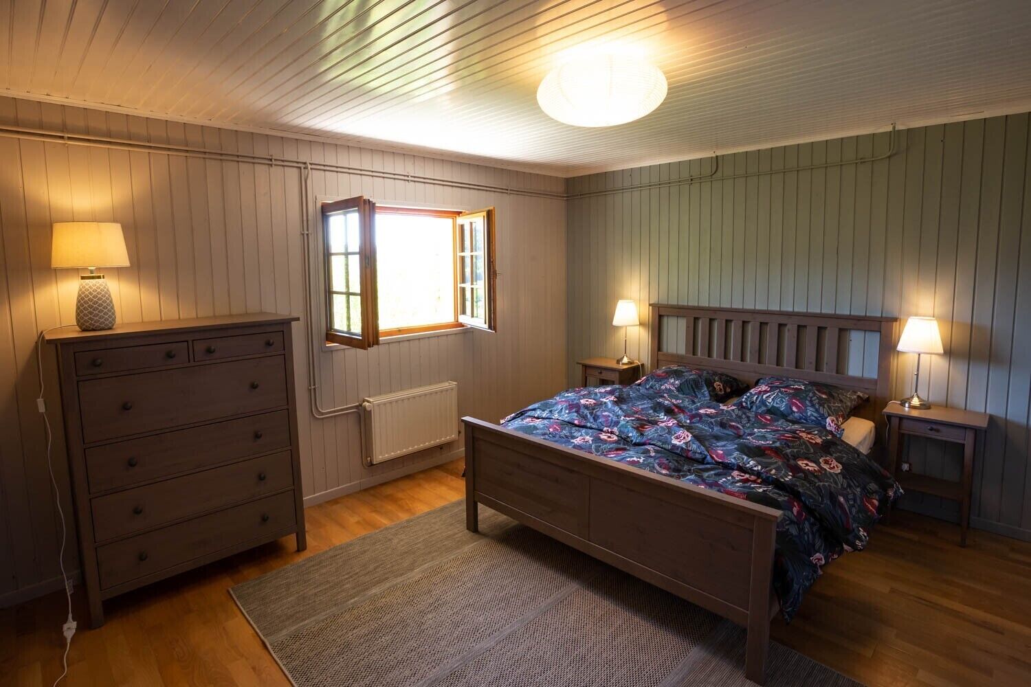 A bedroom with a wooden bed, dresser, and nightstands, featuring a window and a radiator.