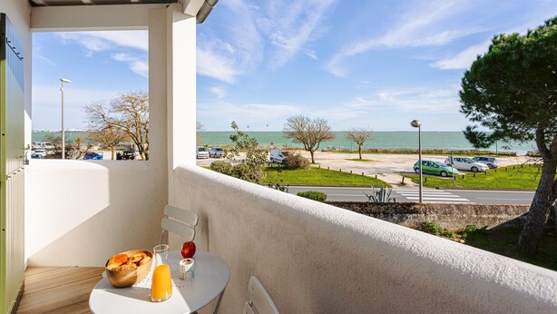 Un balcon avec une table, une chaise et une vue sur la mer et le parking.