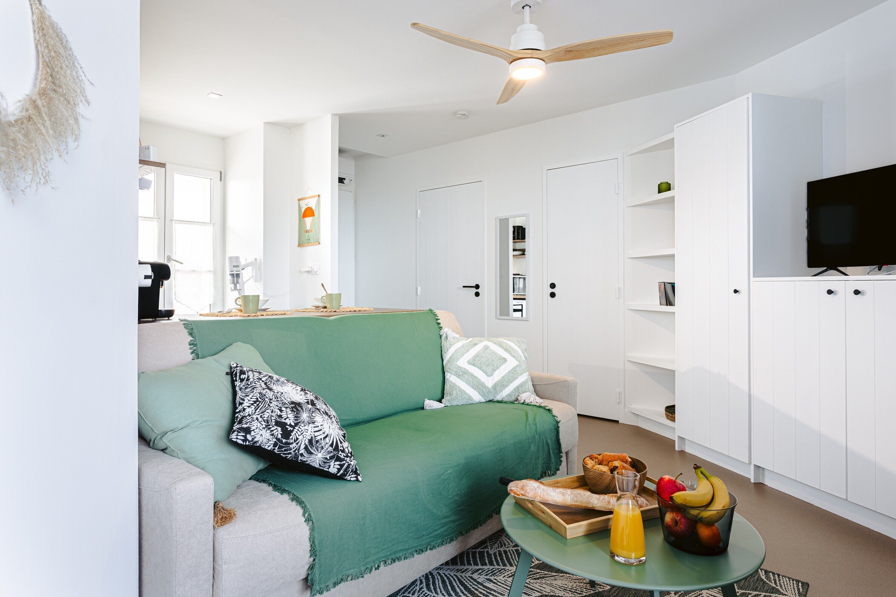 A modern living room with a green sofa, a ceiling fan, a flat-screen TV, and a kitchen area with cabinets and a fruit bowl.