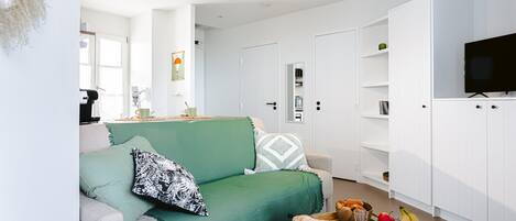 A modern living room with a green sofa, a ceiling fan, a flat-screen TV, and a kitchen area with cabinets and a fruit bowl.