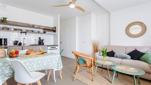 A modern kitchen with white cabinets, a dining table with a green floral tablecloth, and a wicker chair. - La Plage Apartment: Sea View, Private Terrace, Wi-Fi (RIVEDOUX PLAGE)