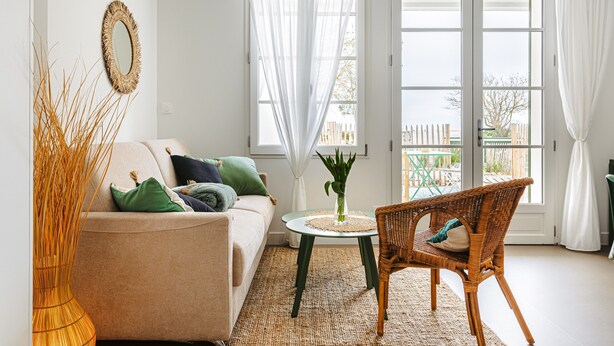 A living room with a sofa, a wicker chair, a round table with a vase, and a view through large windows. - La Plage Apartment: Sea View, Private Terrace, Wi-Fi (RIVEDOUX PLAGE)
