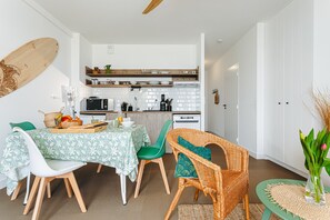 Une cuisine compacte avec des meubles blancs, un four à micro-ondes et une machine à café. On y trouve un coin repas avec une table pour deux personnes, des chaises vertes et un fauteuil en osier.