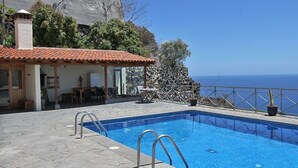 Una piscina con escaleras de metal, un patio con mesas y sillas, y vistas al mar.