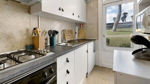 Une cuisine équipée d’une cuisinière à gaz, de meubles blancs, d’un lave-vaisselle et d’une fenêtre donnant sur des palmiers.