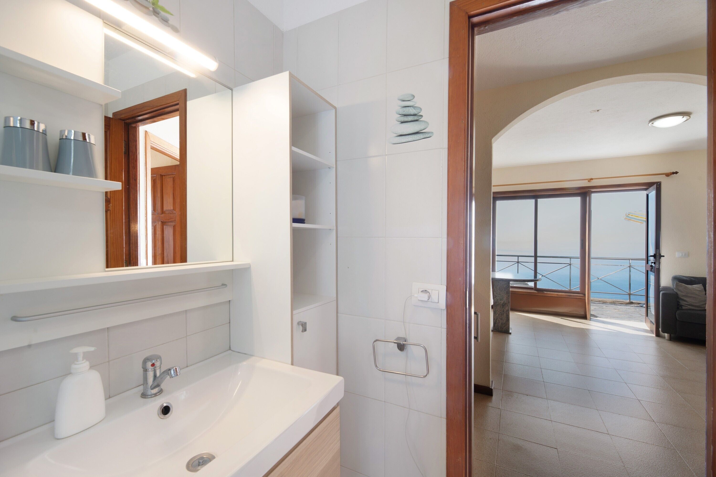 Une salle de bain avec un lavabo, un miroir et des étagères ; une porte donnant sur un balcon avec vue ; et un sol carrelé.