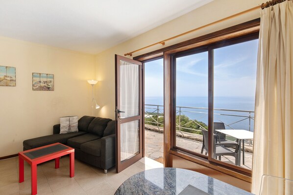 Une pièce comprenant un canapé, une petite table, un balcon avec une table et des chaises, et une vue sur la mer.
