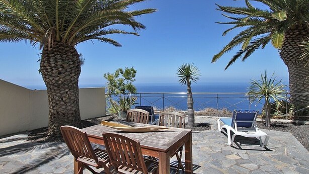 An outdoor patio with a wooden table and chairs, a pool, and palm trees. - Holiday Home 'Moijama 6' with Sea View, Shared Pool and Wi-Fi (Los Llanos de Aridane)