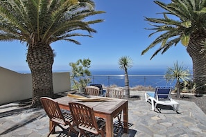 An outdoor patio with a wooden table and chairs, a pool, and palm trees. - Holiday Home 'Moijama 6' with Sea View, Shared Pool and Wi-Fi (Los Llanos de Aridane)