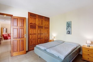 A bedroom with a bed, wooden wardrobe, bedside table, lamp, and a painting on the wall.
