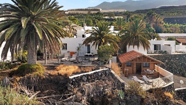 A coastal property with a stone retaining wall, palm trees, and a wooden cabin with outdoor seating. - Apartment 'Moijama 1' with Shared Pool, Private Terrace and Wi-Fi (Los Llanos de Aridane)