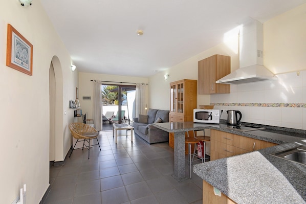A modern kitchen with a microwave, kettle, and granite countertop. There is a dining area with a table and chairs, and a living room with a sofa and a TV.
