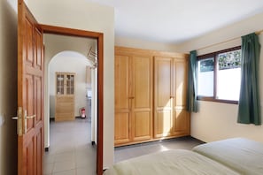A bedroom with a wooden wardrobe, a bed, a window with curtains, and a hallway leading to a kitchen area.