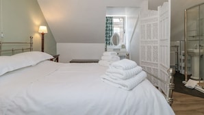 Habitación de hotel con una cama bien hecha, una mesita de noche con lámpara, un baño con espejo y un panel blanco decorativo.