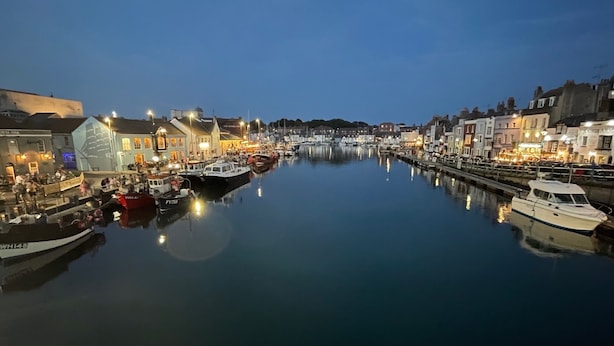 Un puerto deportivo al atardecer con barcos amarrados a lo largo del agua, edificios con ventanas iluminadas y un río tranquilo que refleja las luces.