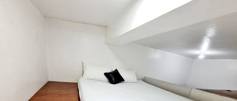 Un dormitorio pequeño y minimalista con una cama individual, paredes blancas y techo inclinado.