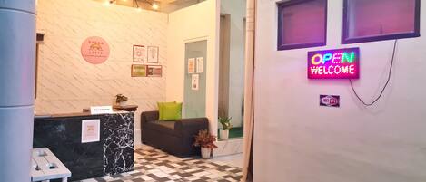 A hotel reception area with a marble reception desk, a sofa with a green pillow, and a neon 'OPEN #WELCOME' sign.