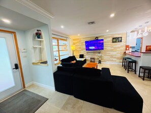 A living room with a black sofa, a TV, a small table, and a kitchen area with stools.