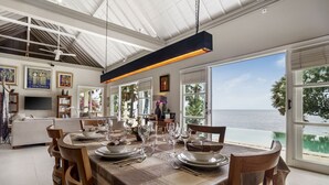 A dining area with a table set for six, large windows with a view of the ocean, and a ceiling fan. - Villa Eden Roc by Alfred in Bali (Karangasem)