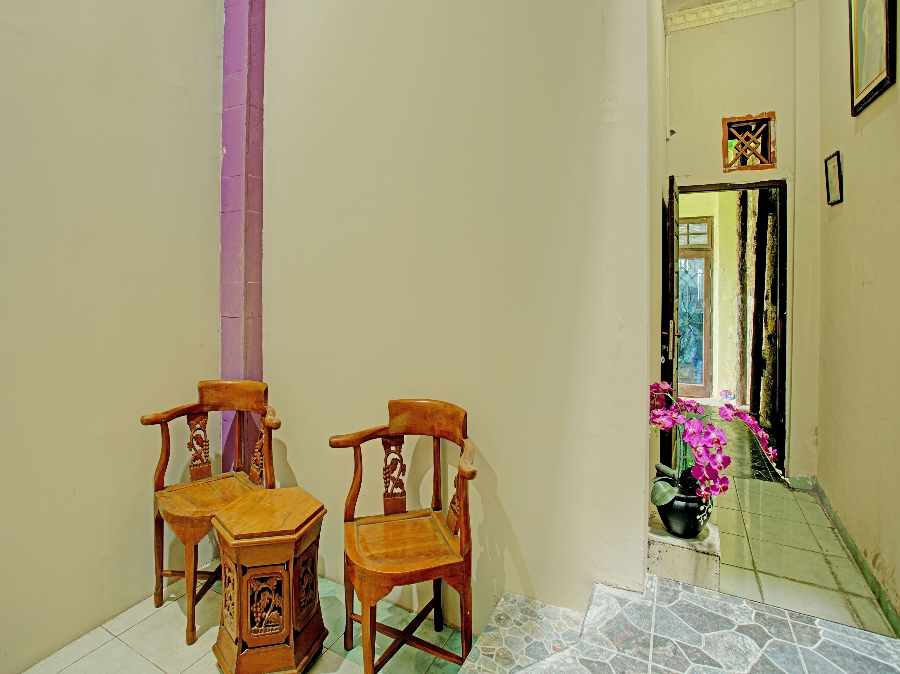 A hallway with two wooden chairs and a small table, a purple pillar, and a potted plant with pink flowers.