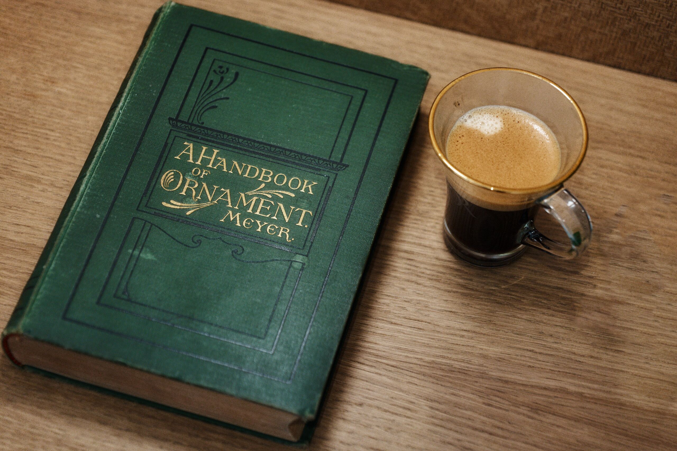 A green book titled "A Handbook of Ornament" next to a glass mug with coffee.