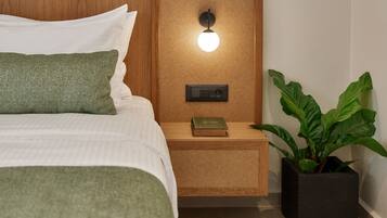 A neatly made bed with white and green pillows, a bedside table with a book and a wall-mounted light, and a potted plant on the floor.