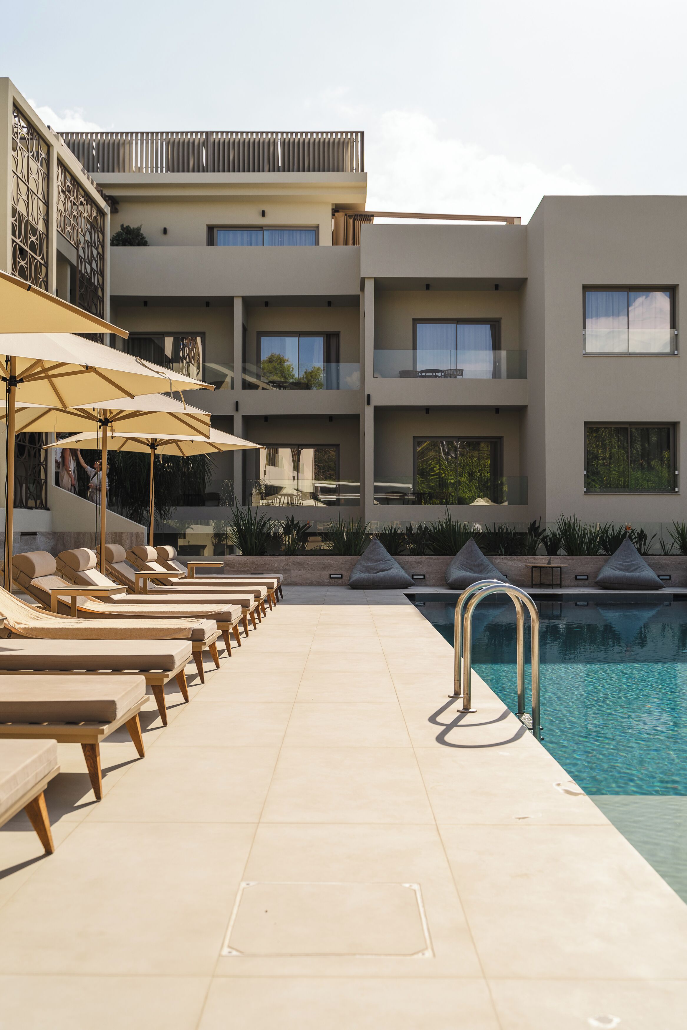A poolside area with sun loungers and umbrellas, a clear pool, and a multi-story building with balconies.