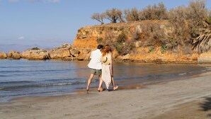 A couple walking along a sandy beach with a rocky cliff and sparse vegetation in the background. - Inea Sole Boutique Hotel (Chania)
