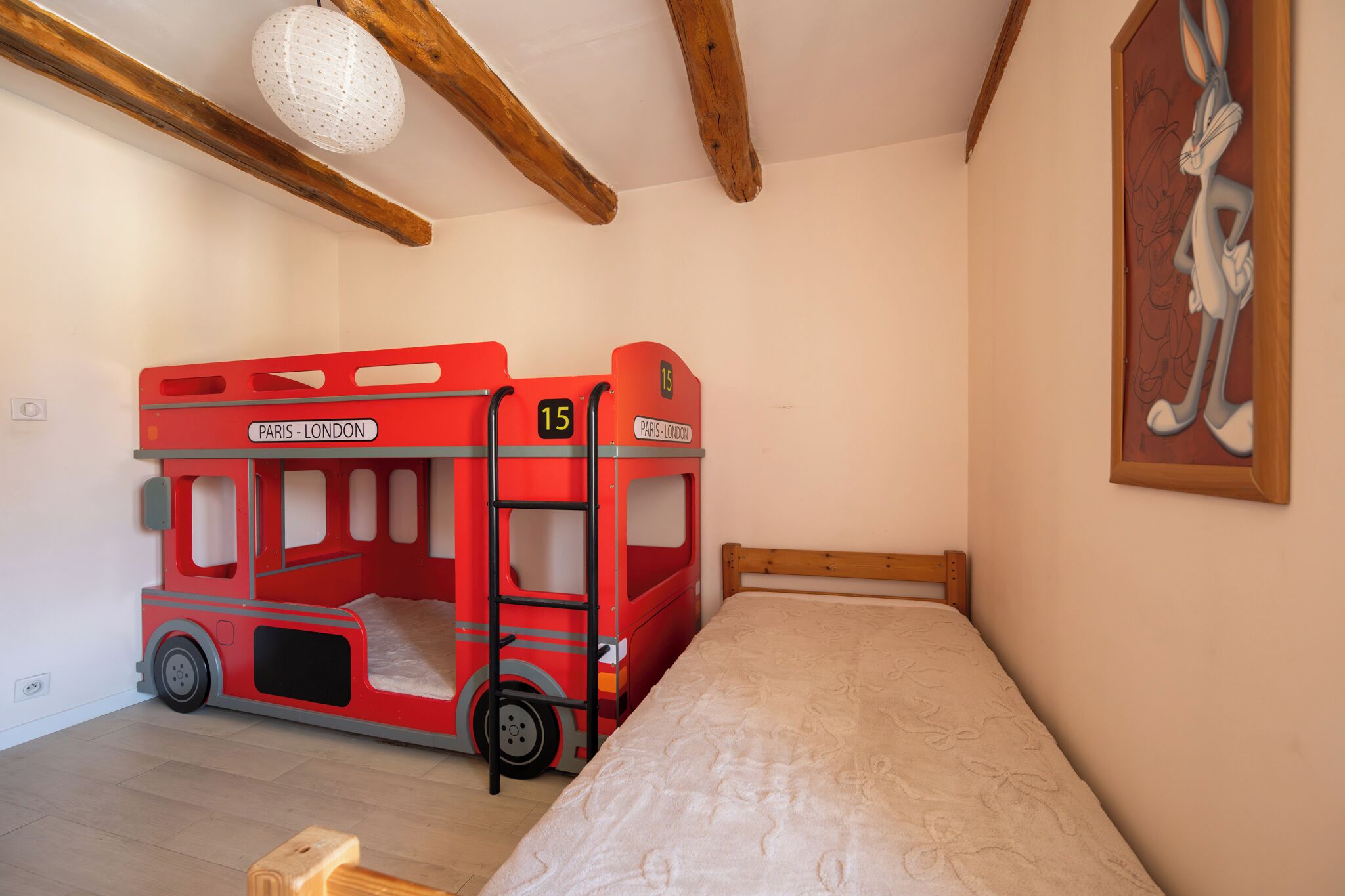 Una habitación con una cama individual, una cama en forma de autobús rojo de dos pisos, vigas de madera, una pared blanca y un personaje de dibujos animados enmarcado.