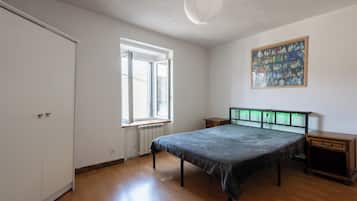 A bedroom with a bed, a radiator, a window, a door, and a nightstand.