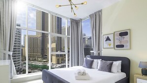 A hotel room with a large window, a bed with pillows, a nightstand with a lamp, and framed artwork on the wall. - LUX  The Continental Marina Suite (Dubai)