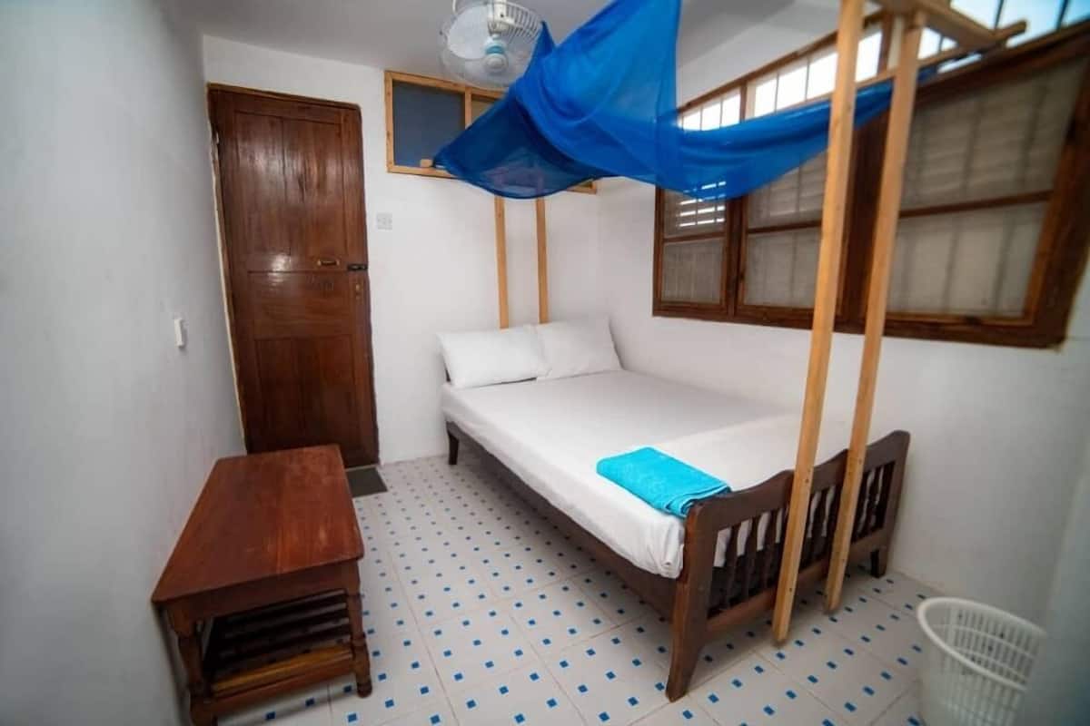 Une petite chambre avec un lit simple, un cadre de lit en bois, une petite table, un ventilateur de plafond et une fenêtre avec des volets en bois.