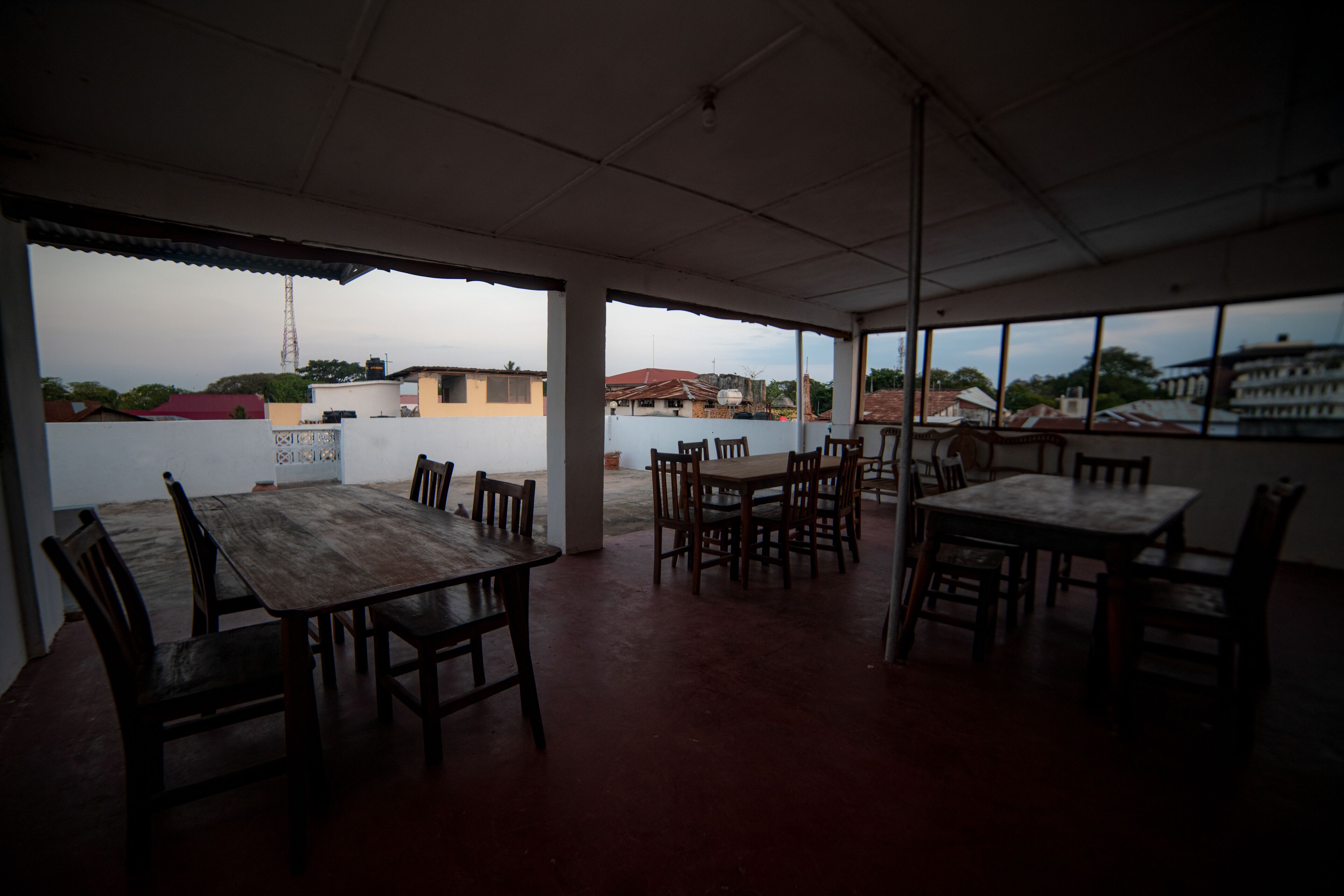 A covered outdoor dining area with multiple wooden tables and chairs, large windows, and a view of buildings in the background.