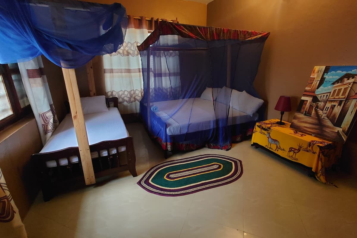Une chambre avec deux lits, dont l’un est à baldaquin et l’autre est simple. On y trouve une petite table avec une lampe et un tableau accroché au mur.