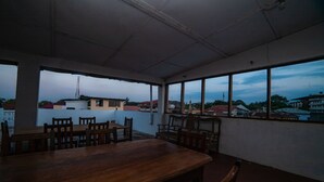 A room with a large wooden table and chairs, large windows with a view of buildings and a tower. - Flamingo Guest House (Zanzibar Town)