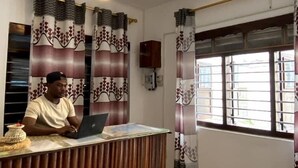 A reception desk with a person working on a laptop, a ceiling fan, patterned curtains, and a window with blinds. - Flamingo Guest House (Zanzibar Town)