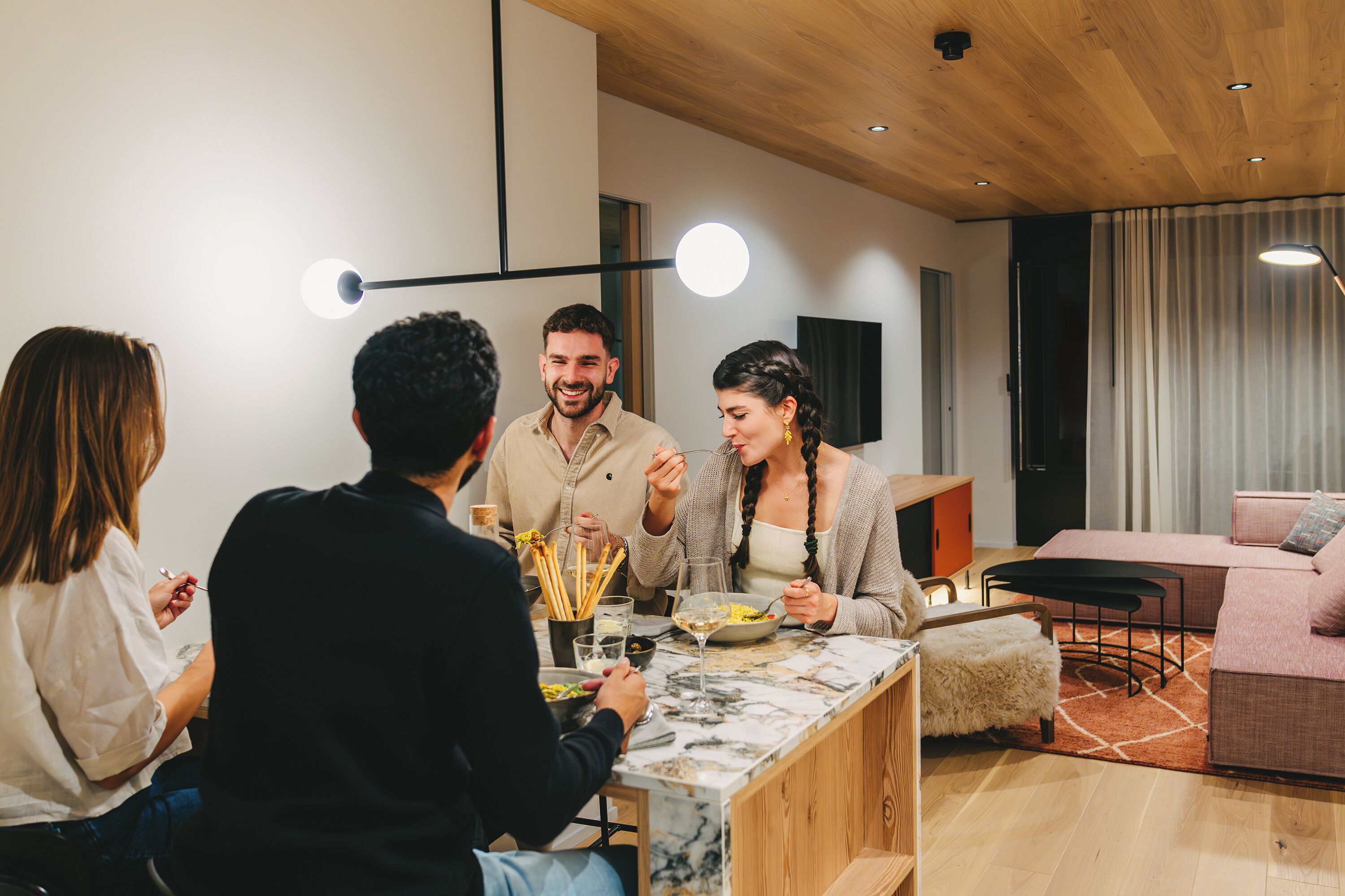 A group of people dining in a modern living room with a marble countertop, wooden furniture, and a large window with curtains.