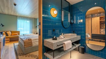 Un dormitorio moderno con cama, mesita de noche y ventana con cortinas. El baño tiene lavamanos, espejo y paredes con azulejos azules.